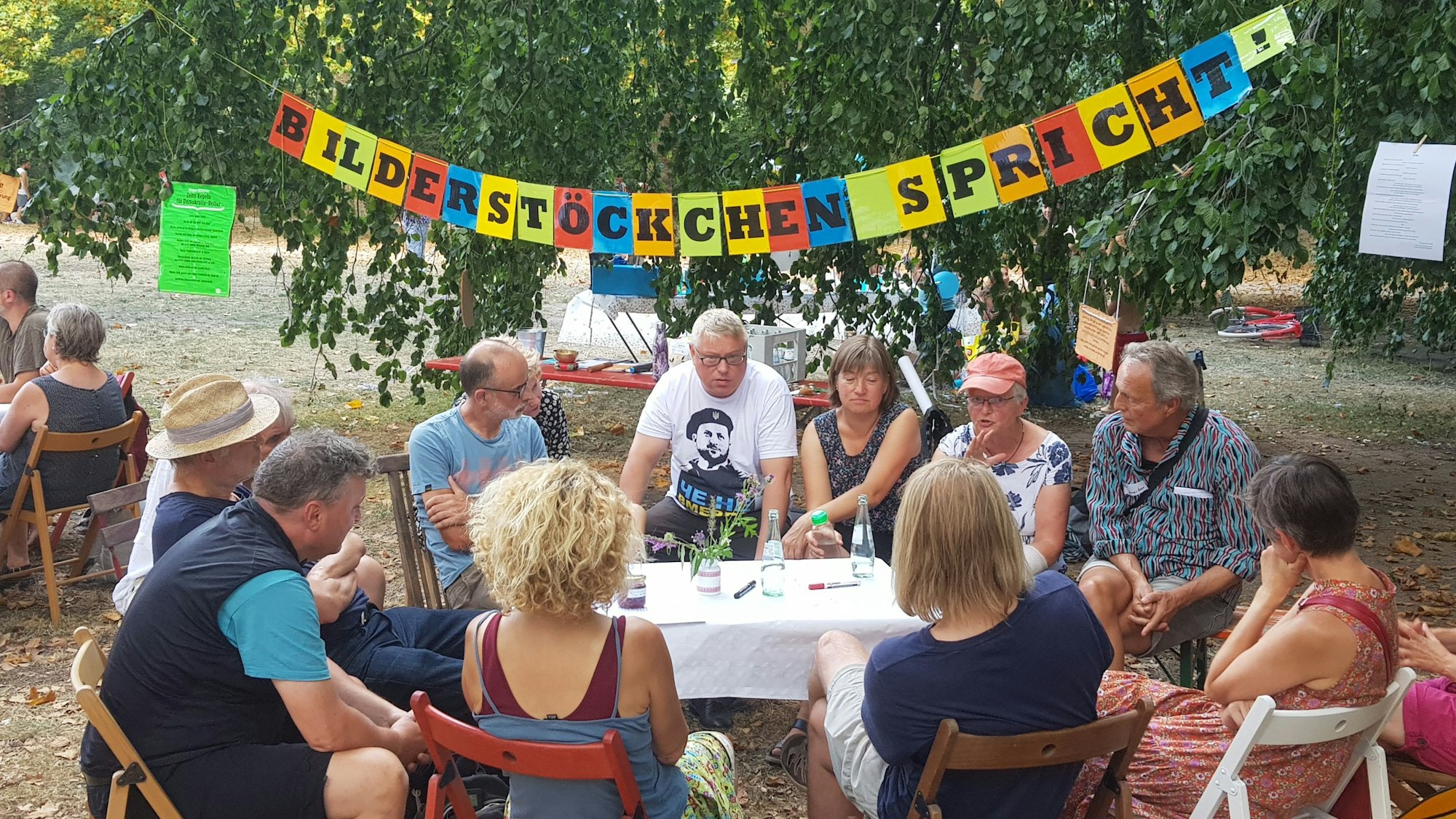 Menschen sitzen im Spätsommer an einem Tisch und diskutieren bei der sechsten Auflage der „Speakers' Corner“ im Blücherpark. Im Hintergrund ist ein Banner mit der Aufschrift „Bilderstöckchen spricht“ zu sehen. Foto von Bernd Schöneck