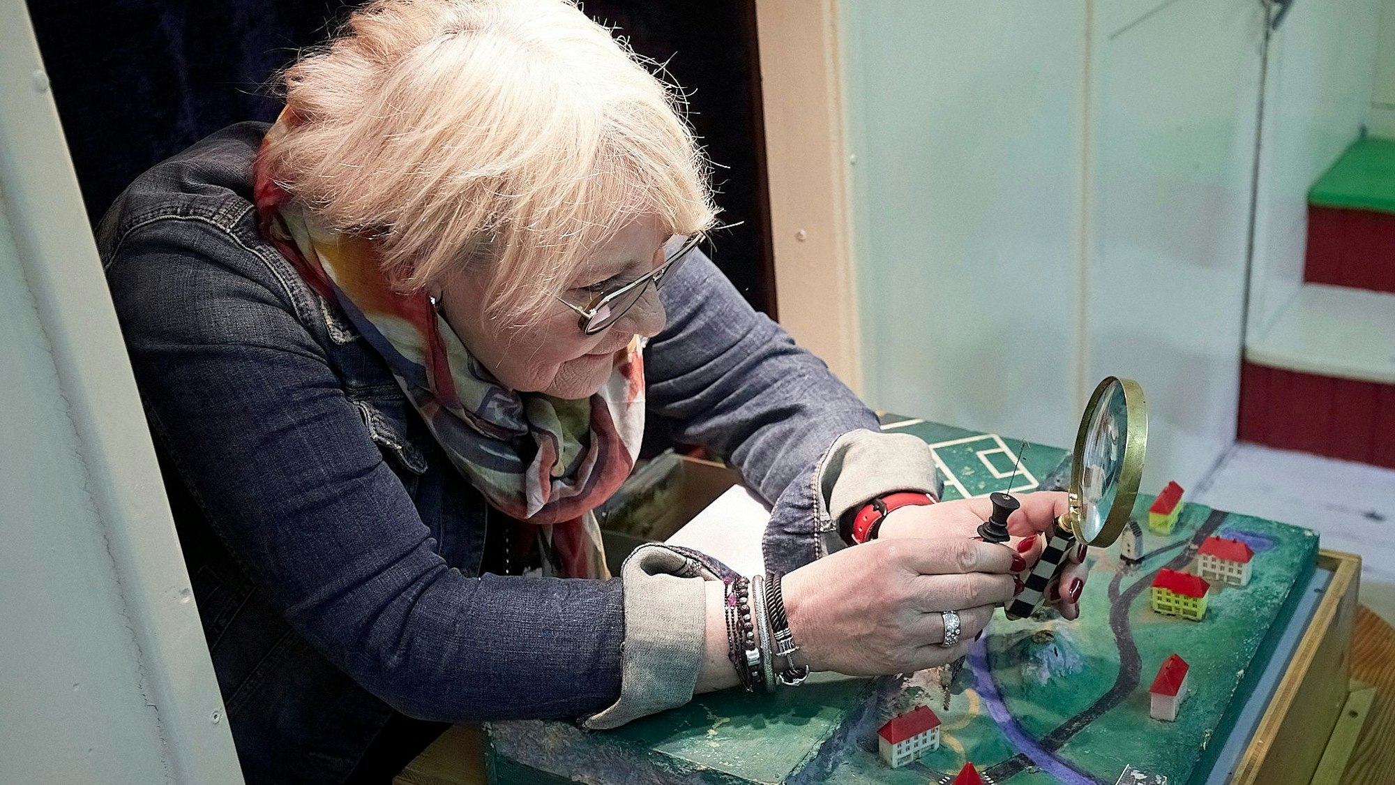 Eine Frau lehnt sich über ein Modellstädtchen und hält eine Lupe in der Hand.