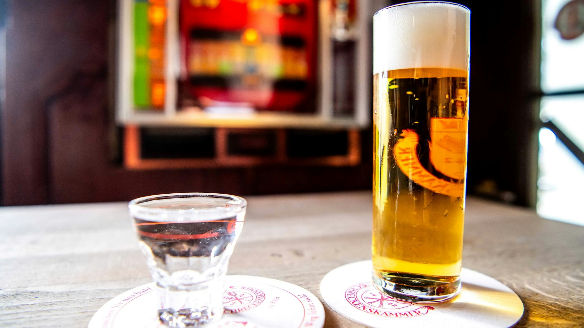 Im Vordergrund stehen ein Kölsch und ein Schnaps, im Hintergrund ist ein Spielautomat zu sehen.