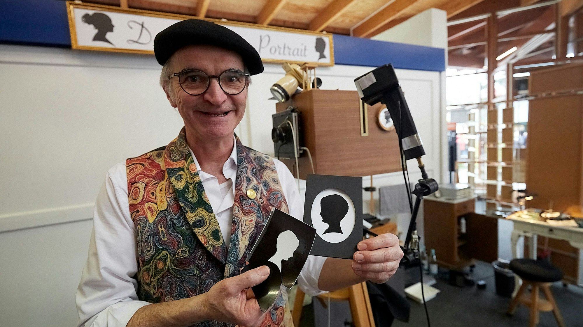 Joachim Zell, in bunter Weste gekleidet, hält zwei Scherenschnitte in der Hand.