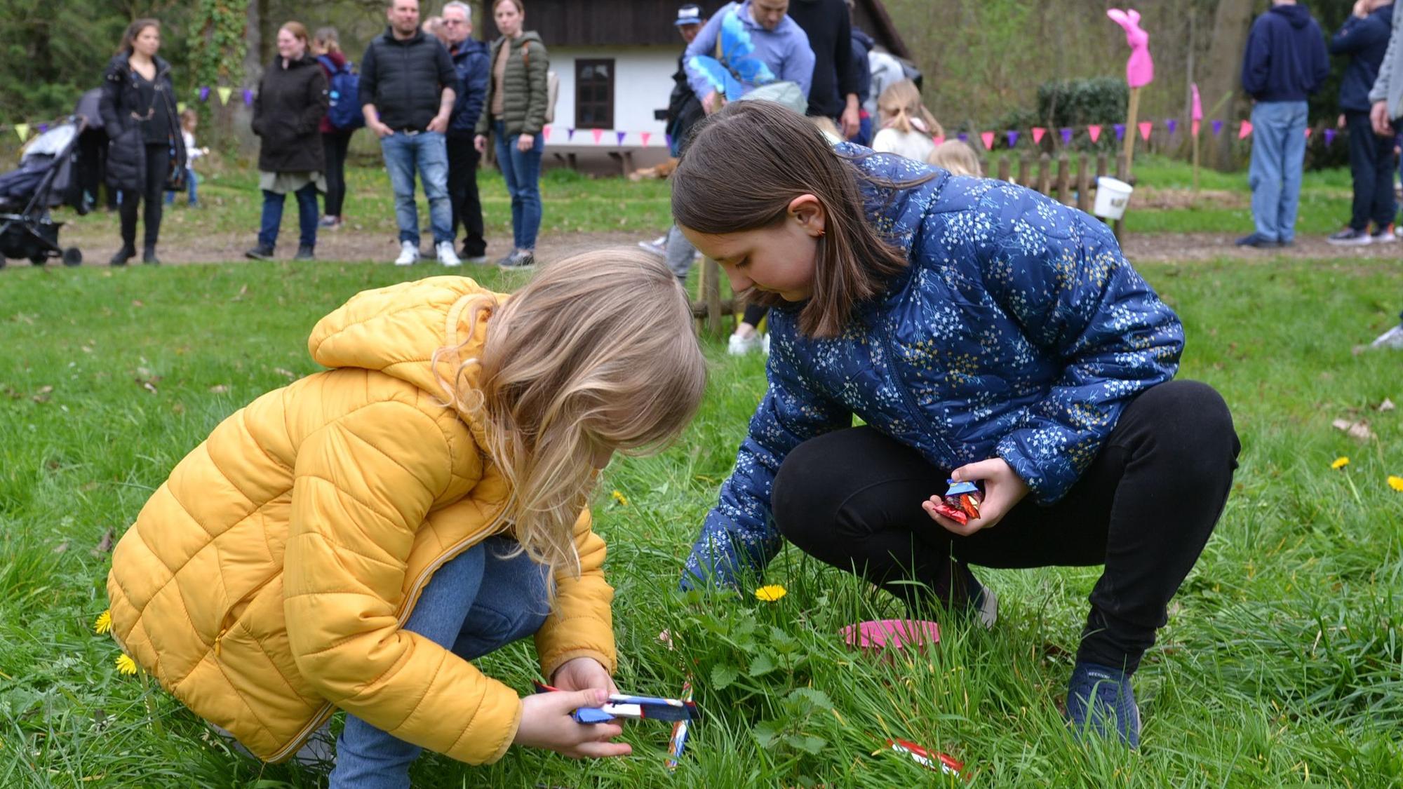 Zwei Kinder suchen im Gras nach Eiern. Im Hintergrund schauen Erwachsene zu.