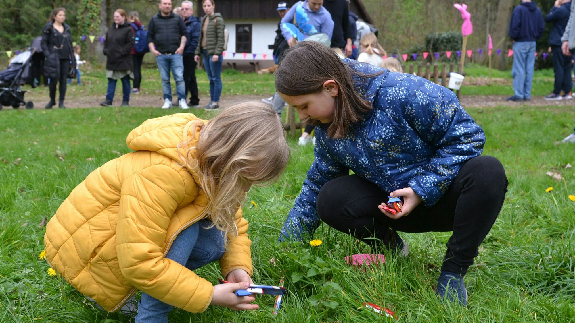 Zwei Kinder suchen im Gras nach Eiern. Im Hintergrund schauen Erwachsene zu.