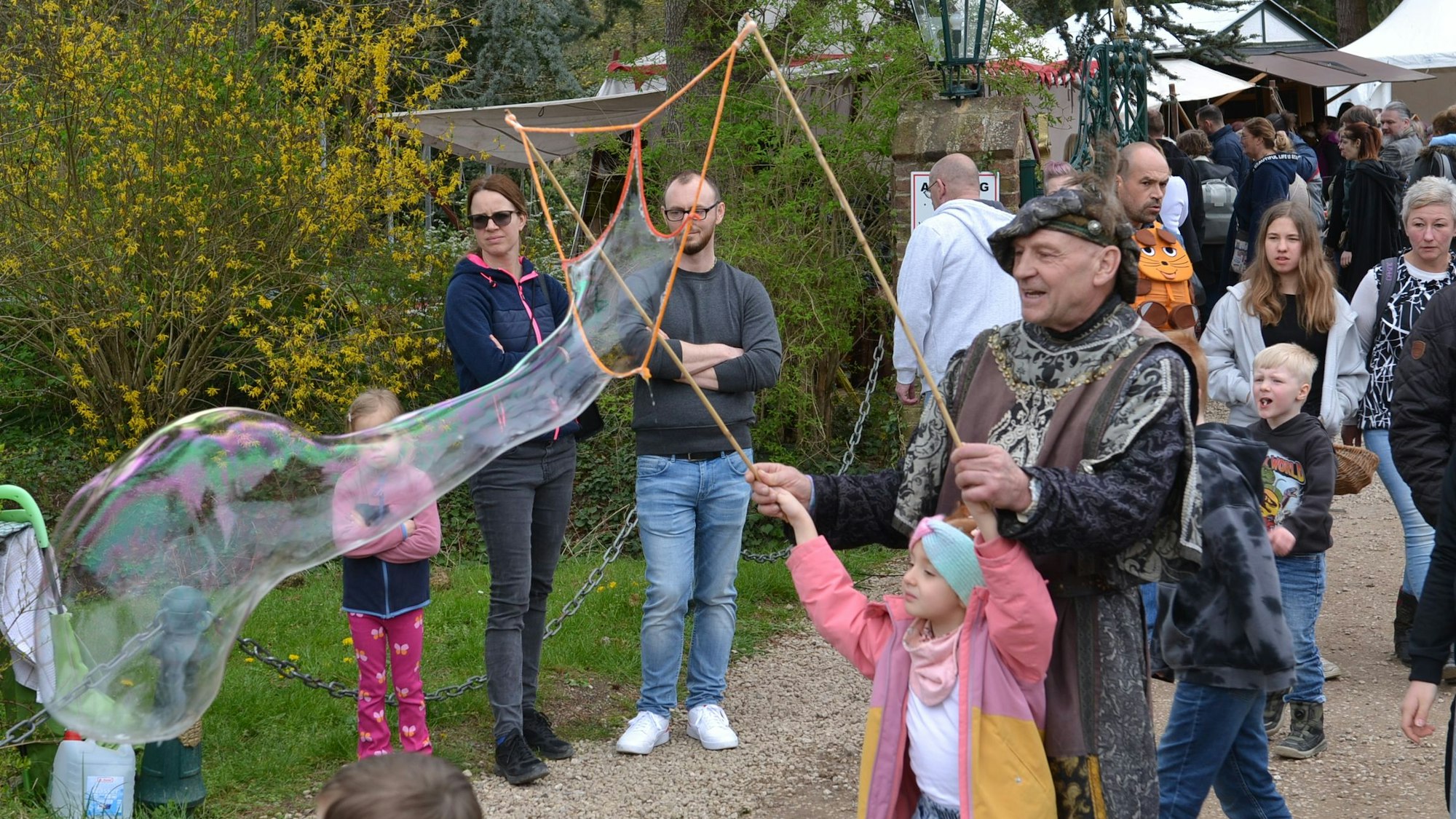 Ein Kind trägt eine große Seifenblase vor sich her, ein mittelalterlich gekleideter Mann hilf ihm dabei.