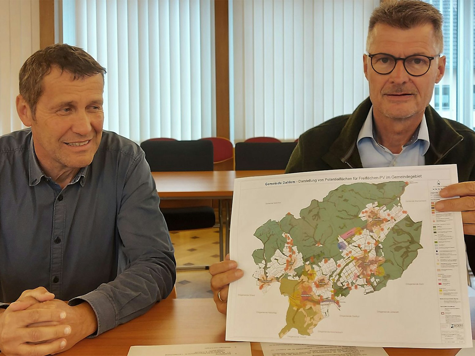 Dahlems Bürgermeister Jan Lembach sitzt an einem Tisch. Er hält eine Karte des Gemeindegebiets in die Kamera. Neben ihm sitzt Erwin Bungartz.