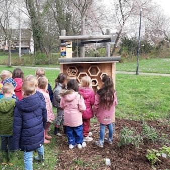 Eine Gruppe von Kindern steht vor dem hölzernen Insektenhotel in der Grünanlage vor der Kita.