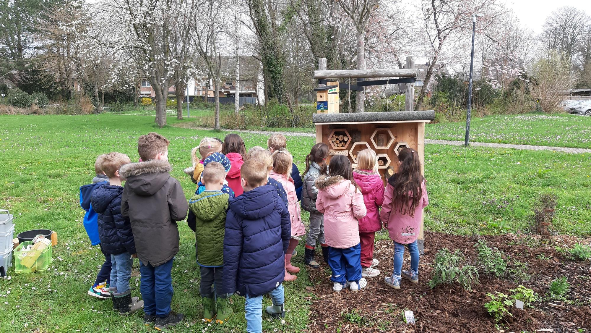 Eine Gruppe von Kindern steht vor dem hölzernen Insektenhotel in der Grünanlage vor der Kita.