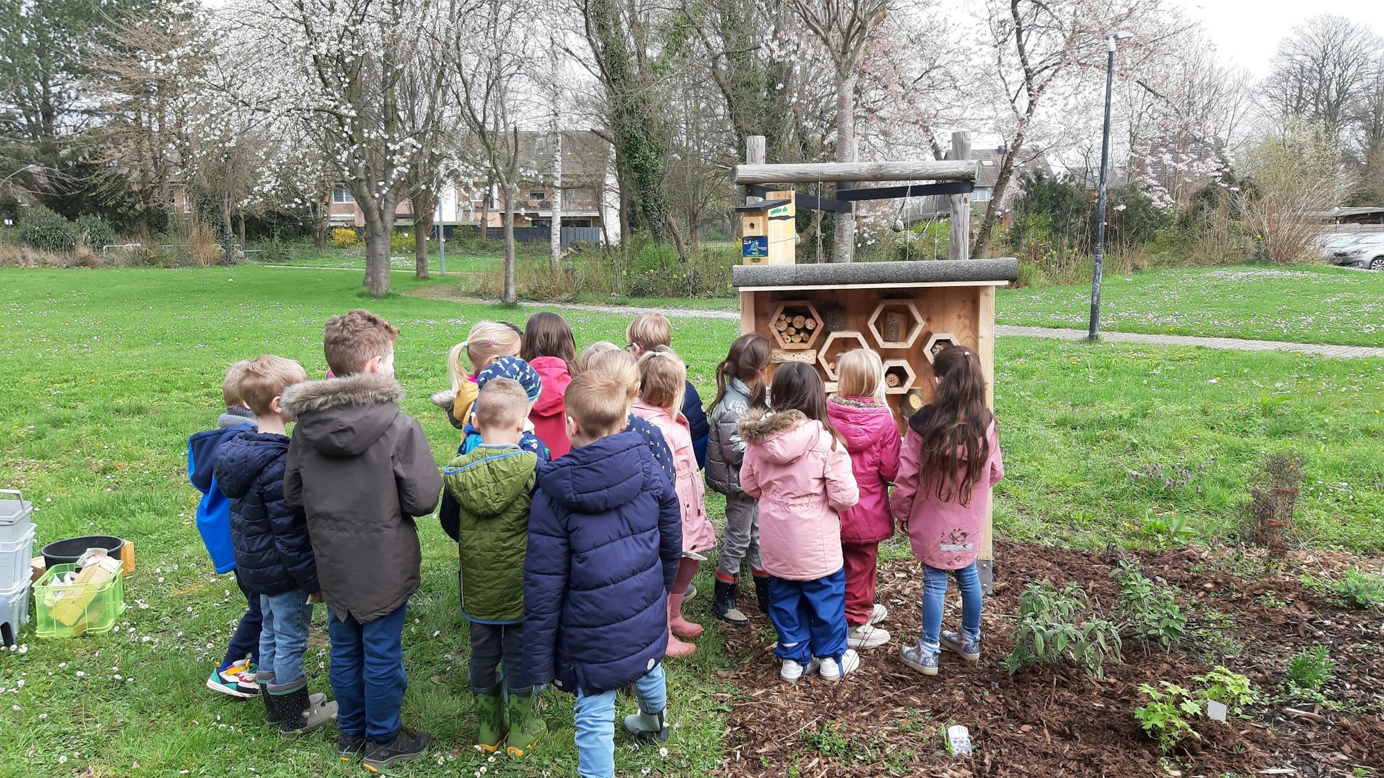 Eine Gruppe von Kindern steht vor dem hölzernen Insektenhotel in der Grünanlage vor der Kita.