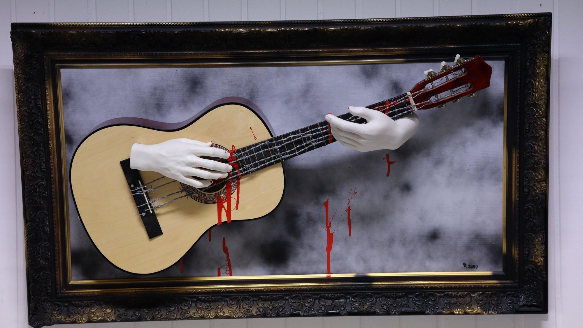 In einem Bilderrahmen spielen blutende Hände eine Gitarre, die mit Stacheldraht bespannt ist.