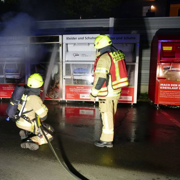 Zwei Feuerwehrleute vor einem brennenden Altkleidercontainer. Eine Person kniet und richtet den Wasserstrahl auf den Sammelbehälter.
