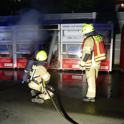 Zwei Feuerwehrleute vor einem brennenden Altkleidercontainer. Eine Person kniet und richtet den Wasserstrahl auf den Sammelbehälter.