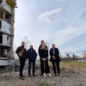 Das Foto zeigt ein Gruppenbild vor einem schwer beschädigten Wohnblock.