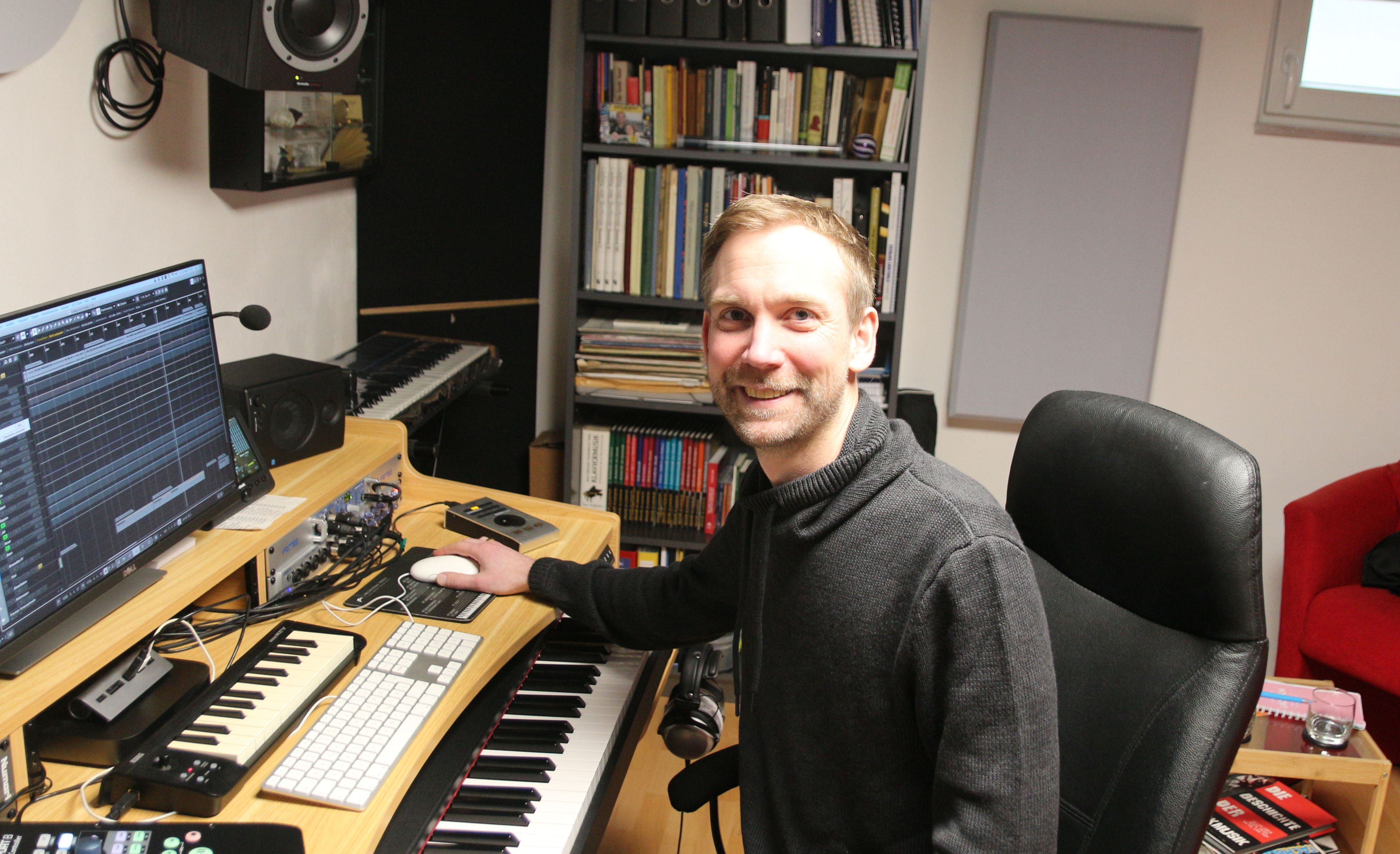 Der Musiker sitzt in seinem Tonstudio und lächelt in die Kamera.