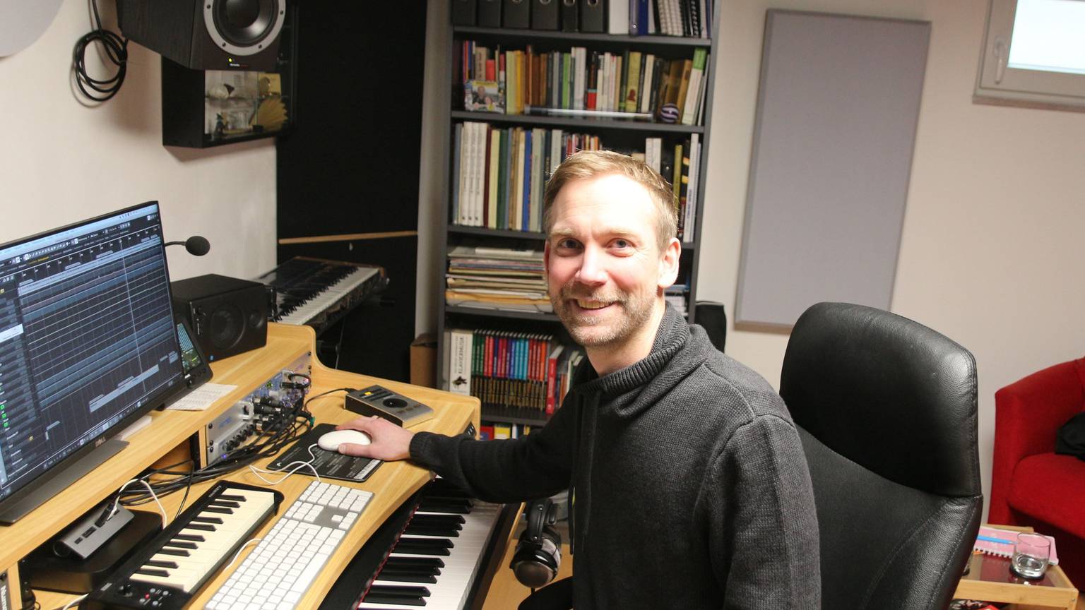 Der Musiker sitzt in seinem Tonstudio und lächelt in die Kamera.