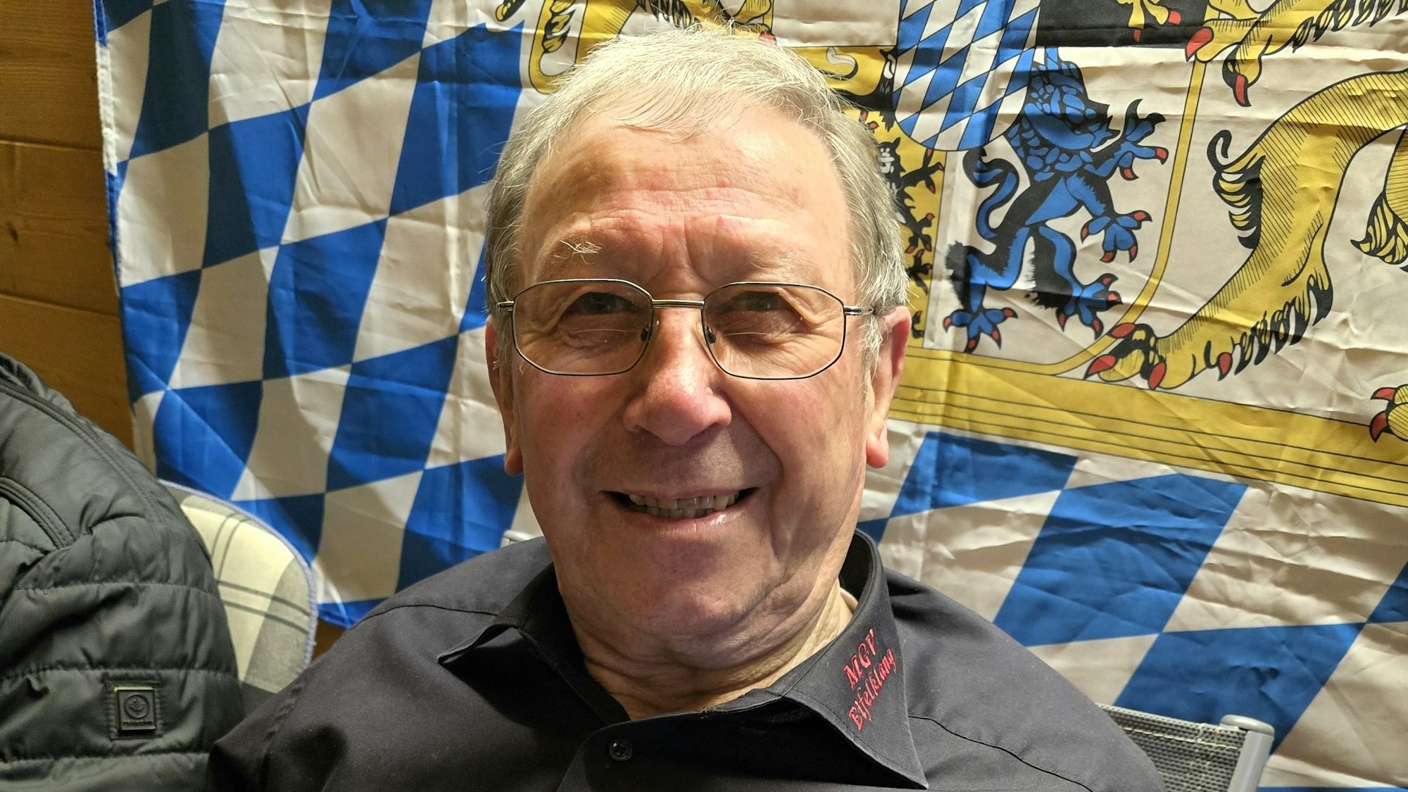 Ewald Weber sitzt vor einer Bayern-Fahne und lacht in die Kamera.
