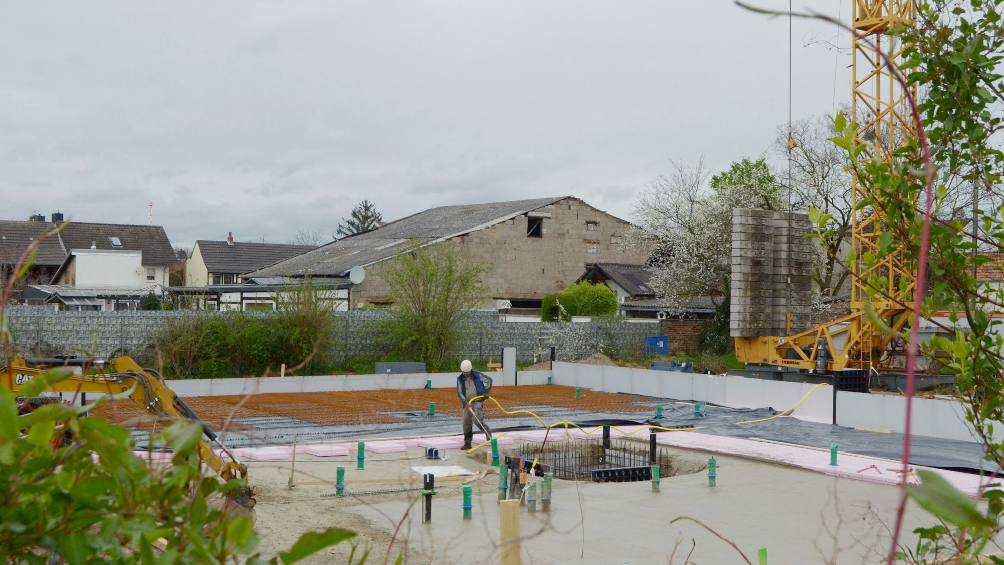 Die Bodenplatte des Neubaus ist bereits gegossen worden. In der Mitte ist ein Bauarbeiter zu sehen, im Hintergrund Wohnhäuser und andere Gebäude.