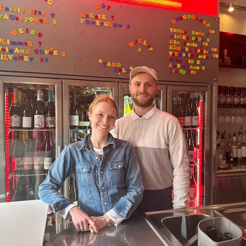 Ein junger Mann und eine junge Frau stehen hinter einer Bar und lächeln
