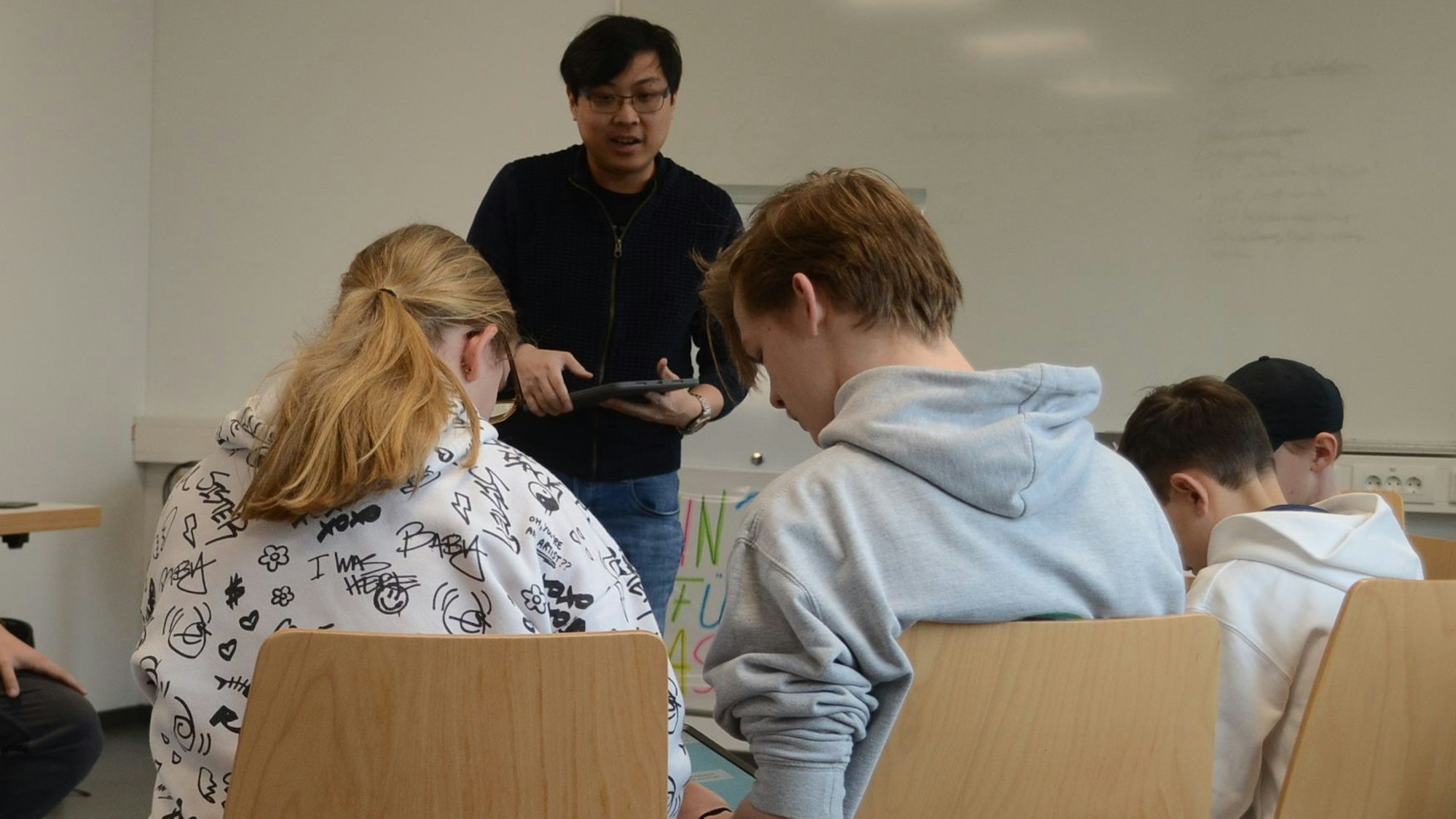 Kinder und Jugendliche beugen sich über Tablet-Computer auf ihrem Schoß. Ein Dozent steht in der Mitte.