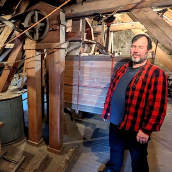 Wolfgang Pütz zeigt die alten Gerätschaften im Dachboden der Hauserbachmühle. Er trägt ein rot-kariertes Holzfällerhemd.