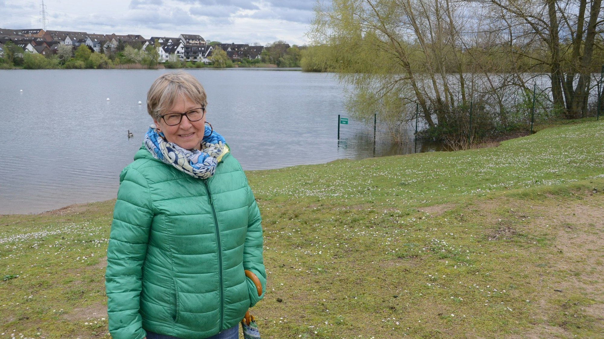 Eine Frau mit grüner Daunenjacke steht am Ufer eines Sees. Im Hintergrund sind Häuser jenseits des Sees zu sehen.