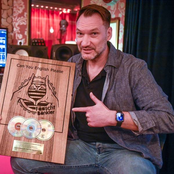 Ein Mann sitzt in einem Tonstudio vor einem Mischpult mit Bildschirm und zeigt eine Holztafel mit drei eingelassenen CDs.
