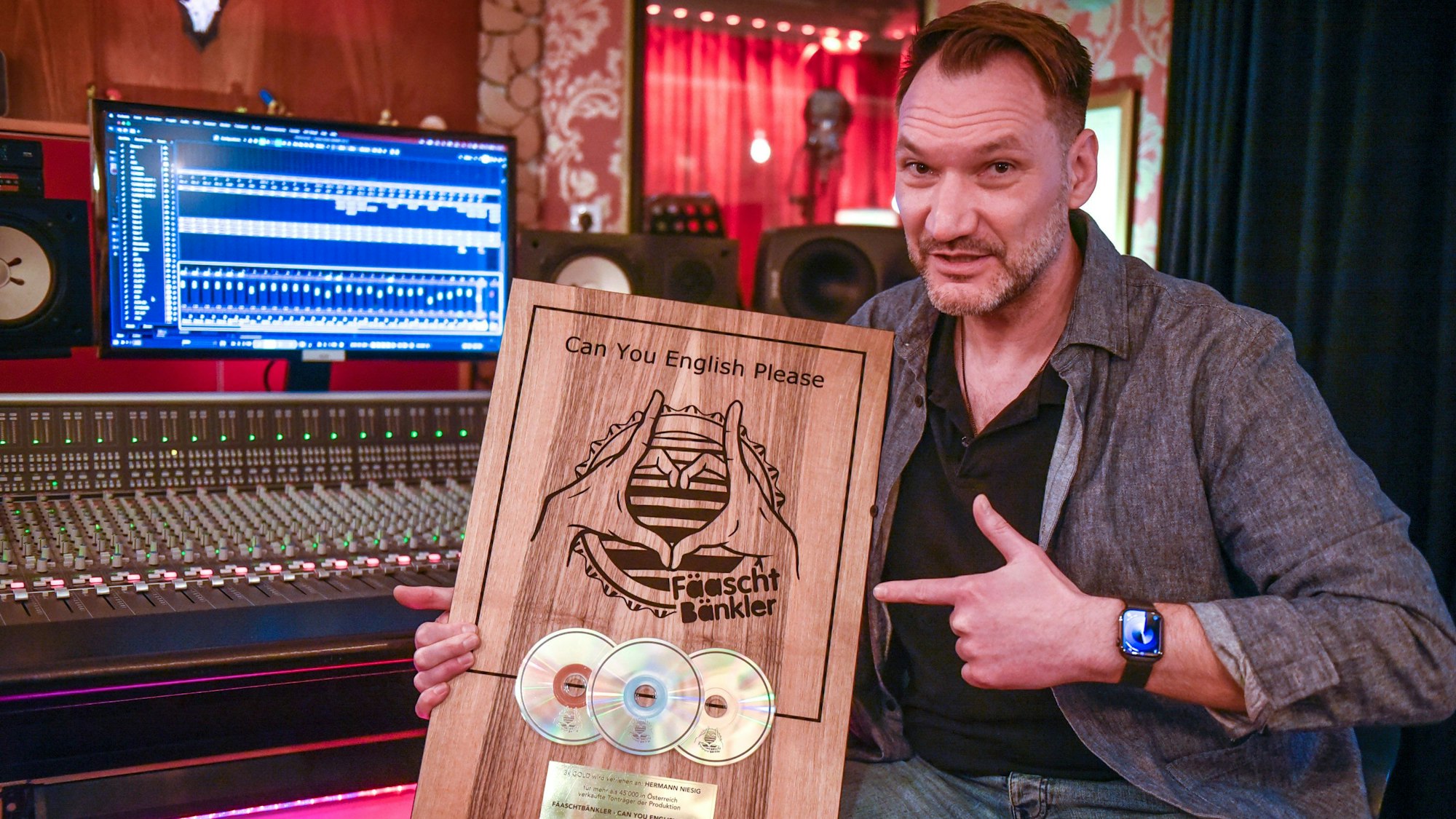 Ein Mann sitzt in einem Tonstudio vor einem Mischpult mit Bildschirm und zeigt eine Holztafel mit drei eingelassenen CDs.