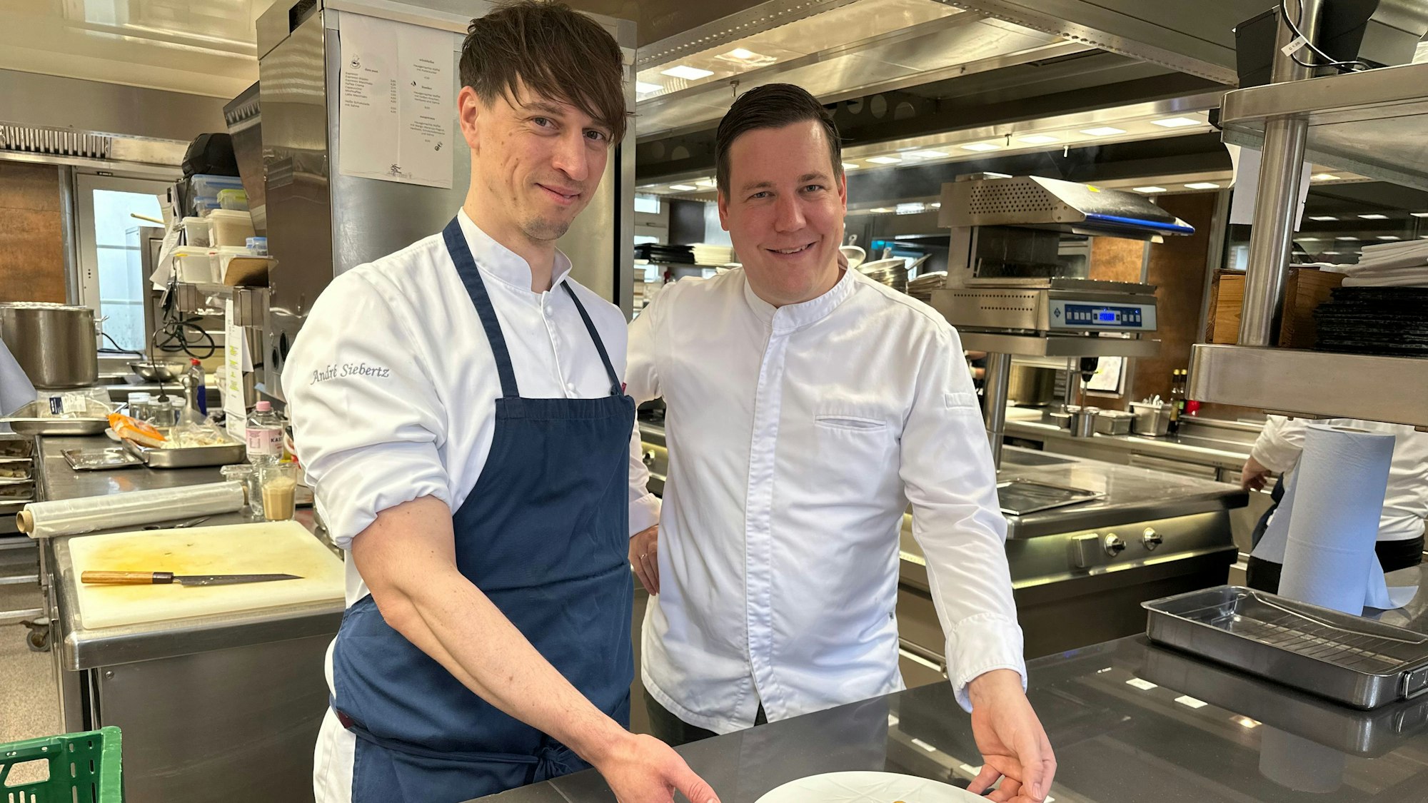 Zwei Männer stehen in einer Restaurant-Küche und zeigen einen elegant angerichtetes Gericht auf einem weißen Teller.