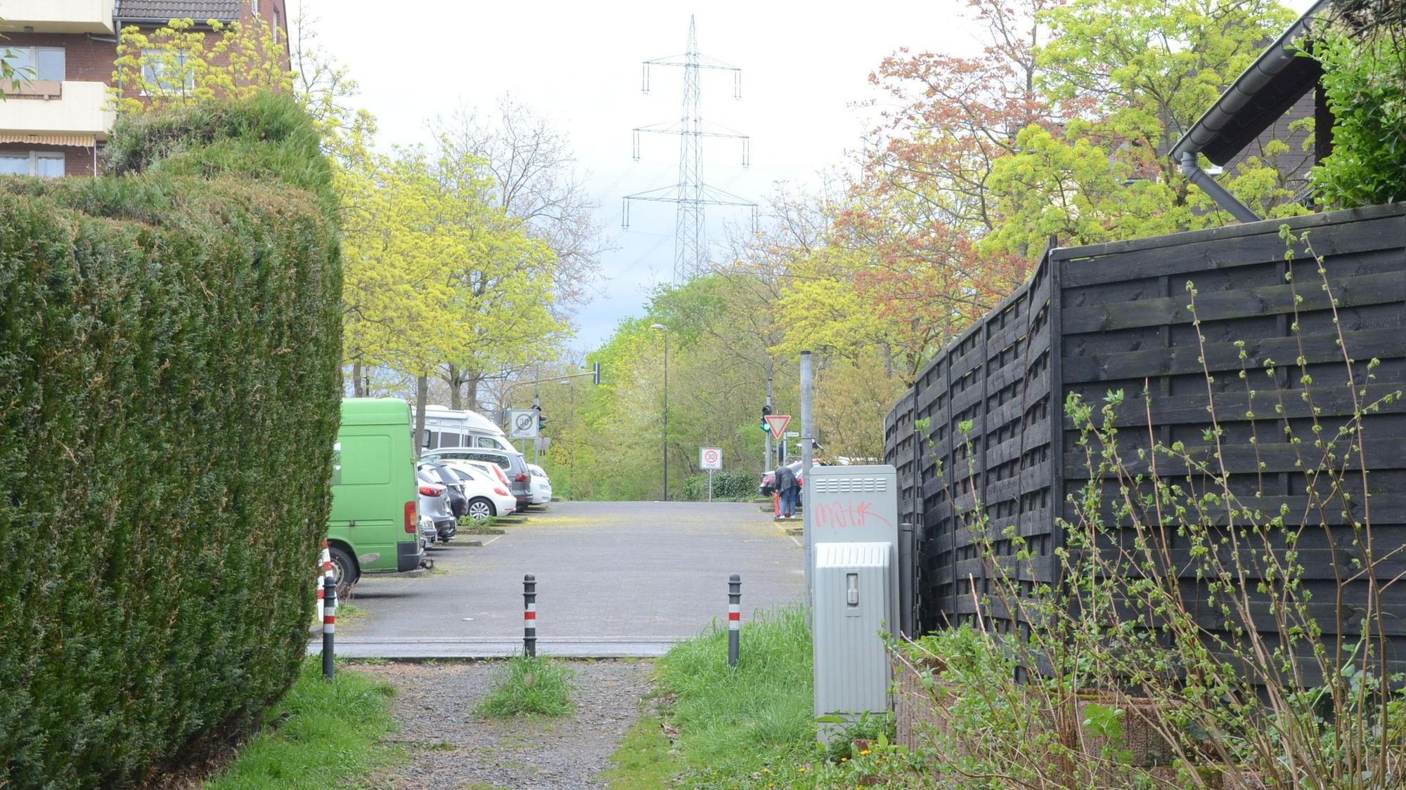 Der Übergang eines Fußwegs zur Straße. Pfosten sperren die Zufahrt für Autos; links wächst eine dichte Hecke, rechts steht ein Zaun.