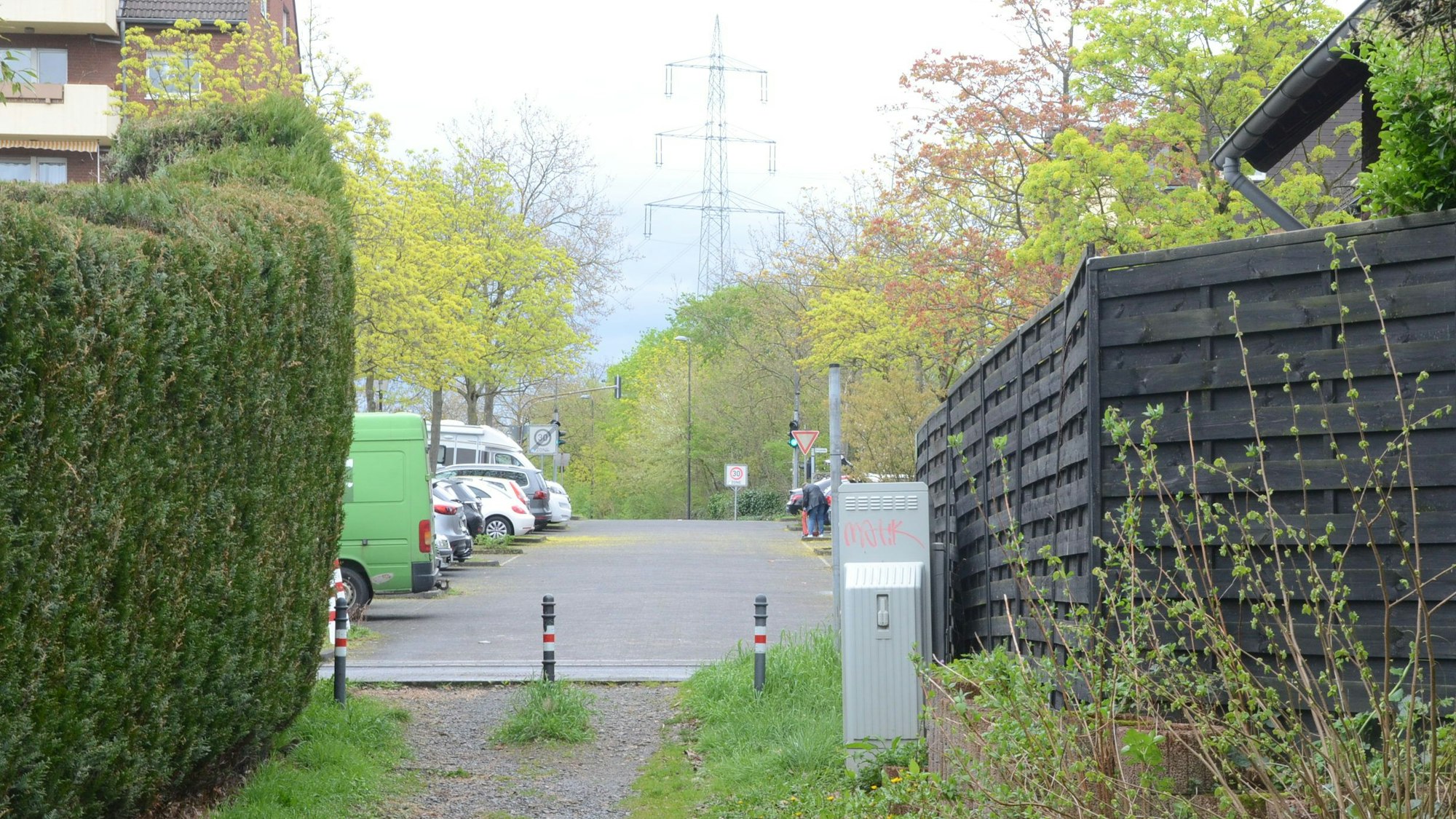 Der Übergang eines Fußwegs zur Straße. Pfosten sperren die Zufahrt für Autos; links wächst eine dichte Hecke, rechts steht ein Zaun.