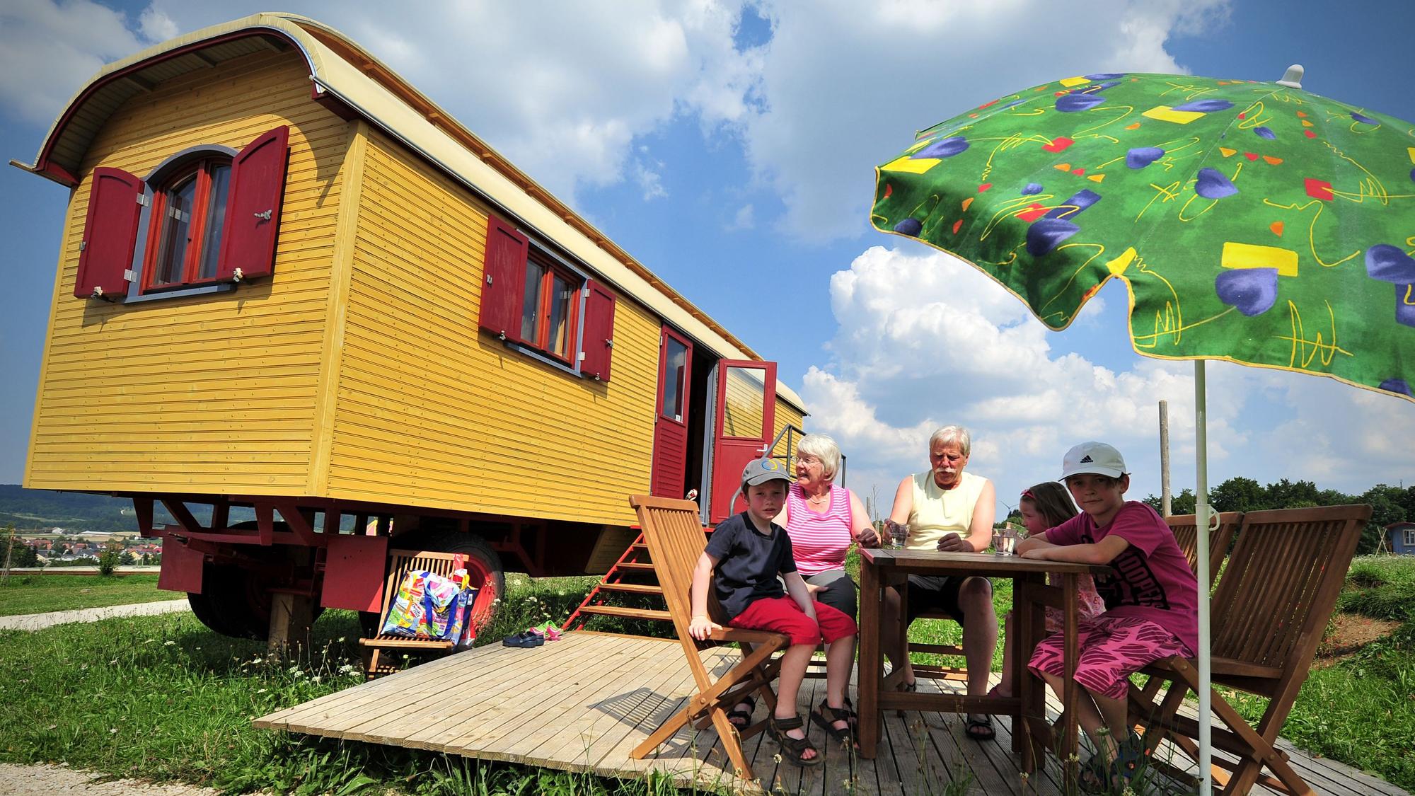 Ein gelber Zirkuswagen, davor sitzen Großeltern mit drei Kindern unter einem Sonnenschirm am Tisch.