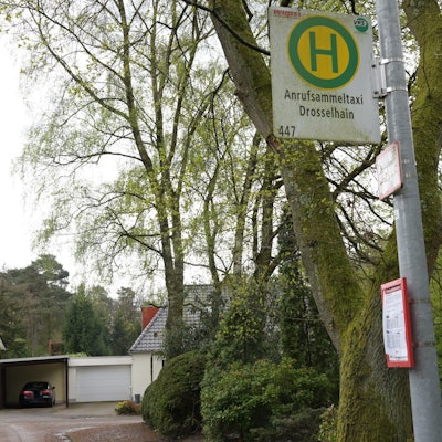 Man sieht das Haltestellenschild, im Hintergrund sind Bäume und Wohnhäuser.