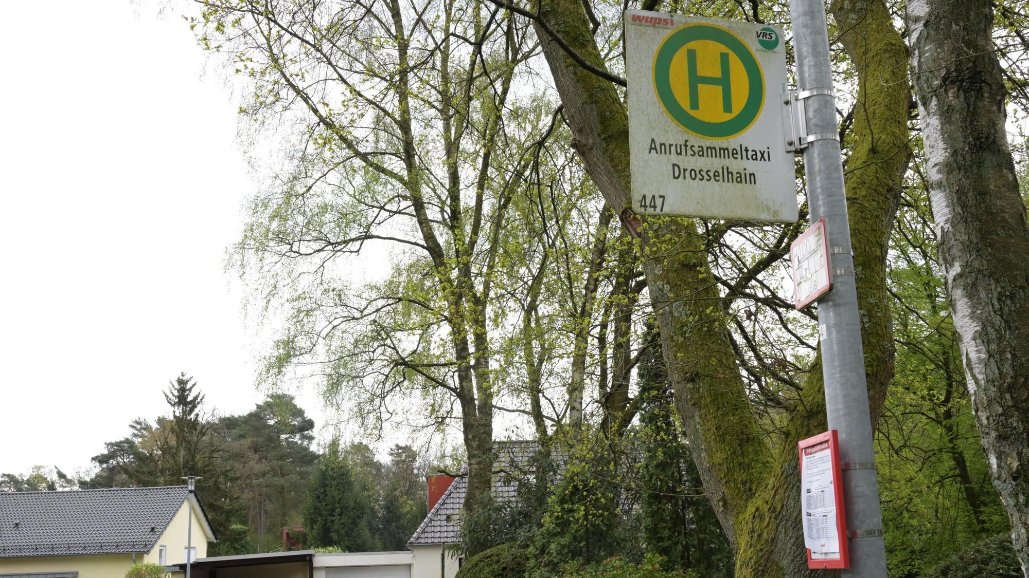 Man sieht das Haltestellenschild, im Hintergrund sind Bäume und Wohnhäuser.