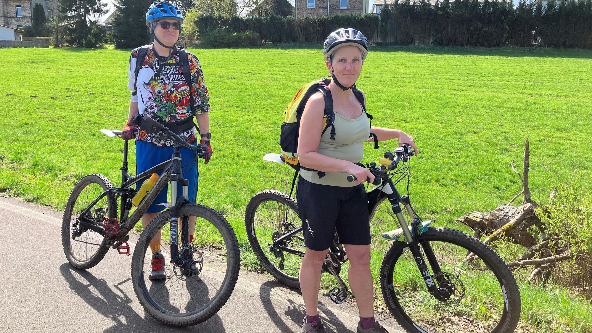 Ein Ehepaar steht mit Mountainbikes an einem Radweg neben einer Wiese.