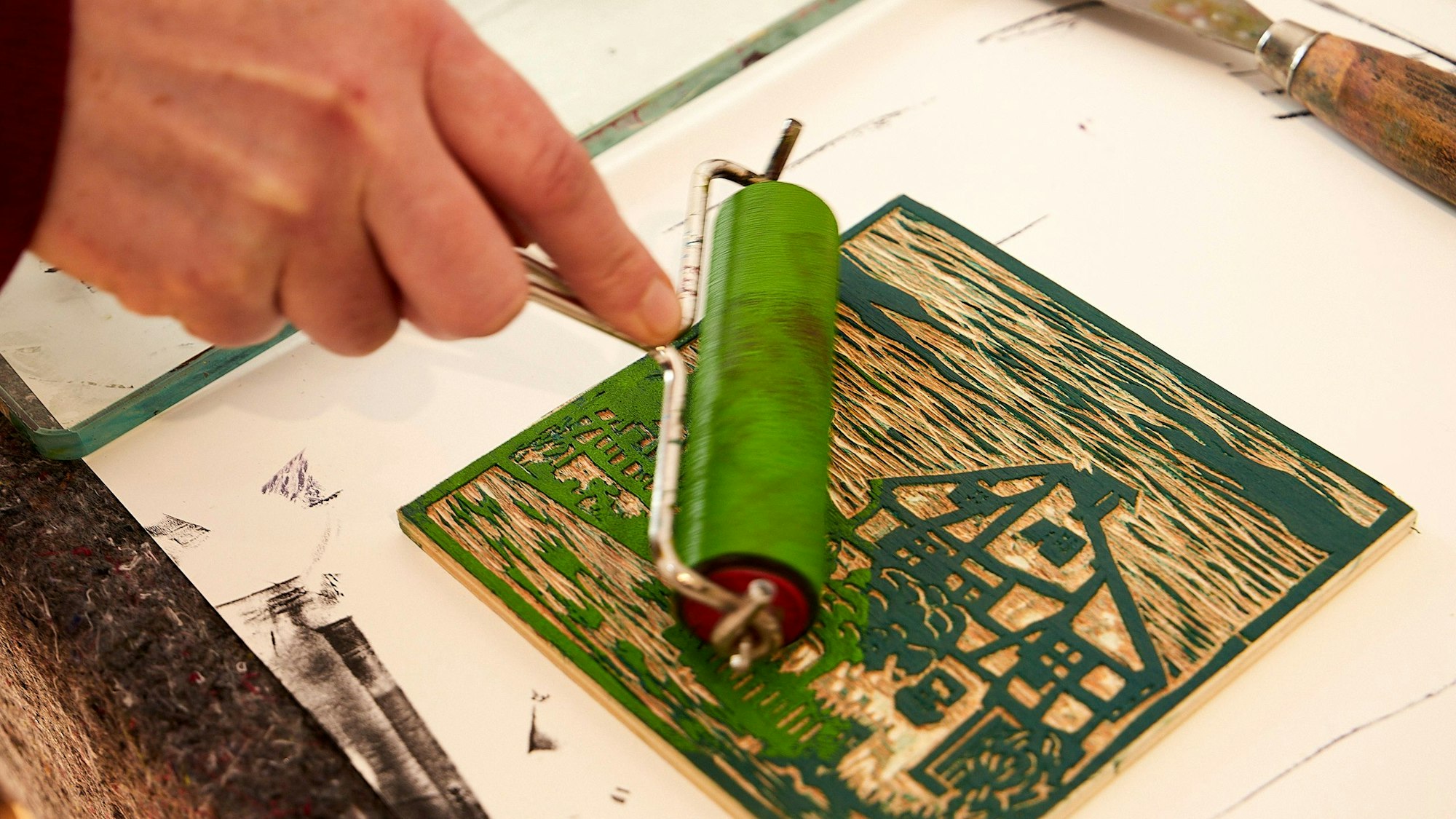 Mit einer Farbrolle wird ein Holzschnitt, der ein Fachwerkhaus in der Natur zeigt, gleichmäßig grün eingefärbt.