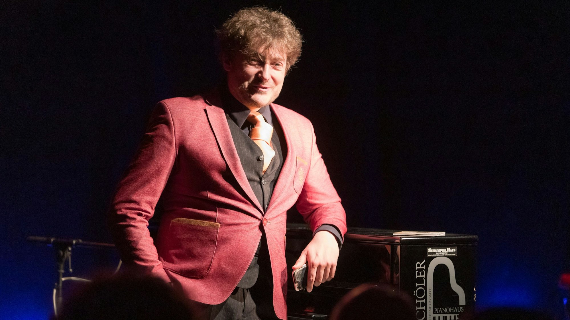 Ein Mann im roten Jackett lehnt an einem Flügel.