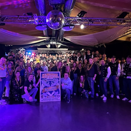 Zwei Chöre stehen im blauen Licht im Saal Altenrath in Marialinden und halten ein Plakat für ein Benefizkonzert.