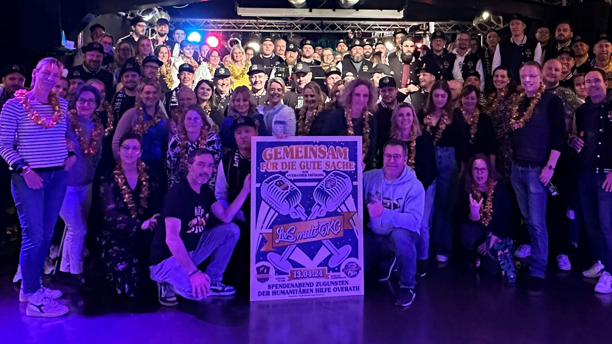 Zwei Chöre stehen im blauen Licht im Saal Altenrath in Marialinden und halten ein Plakat für ein Benefizkonzert.