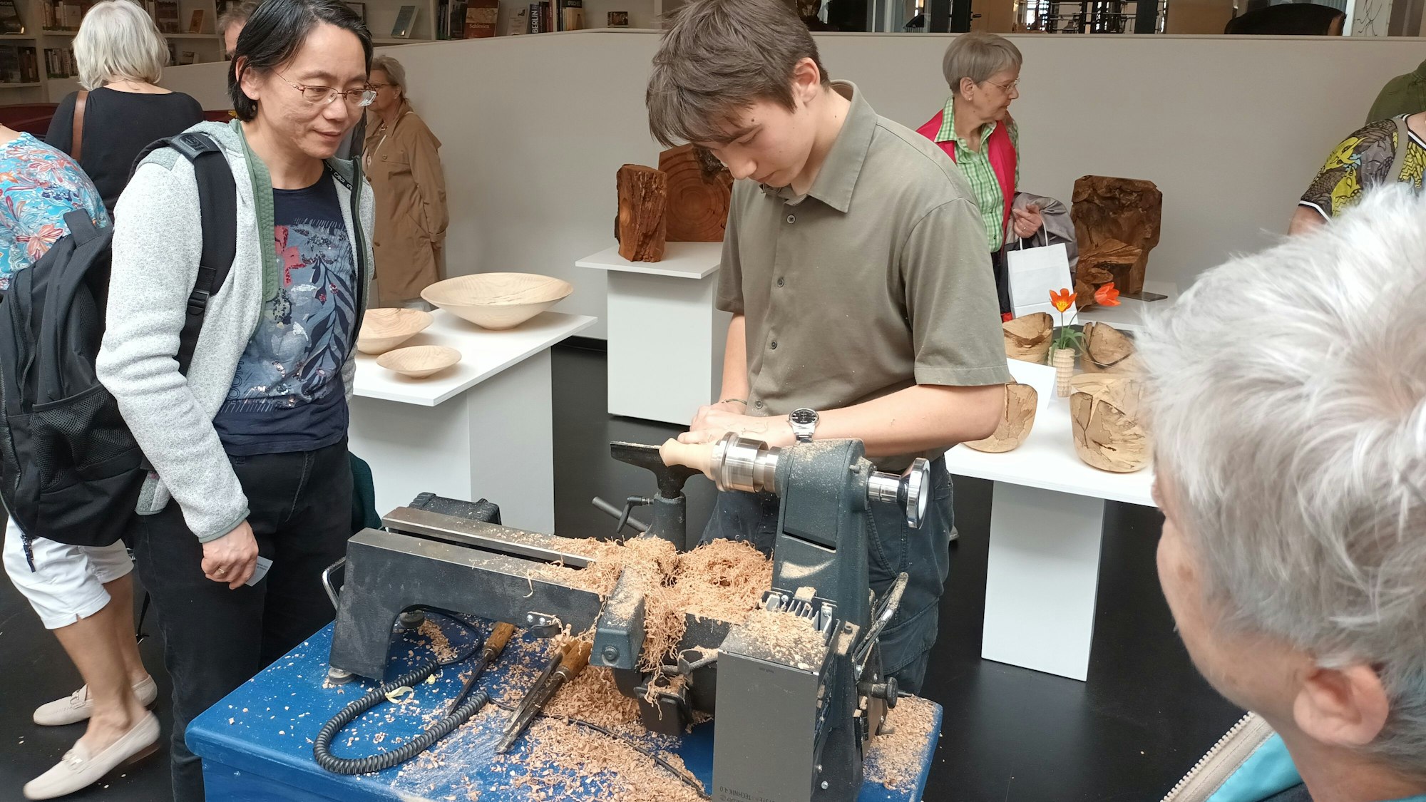 Ein junger Mann steht an einer Holzbearbeitungsmaschine, zwei Frauen gucken ihm zu.