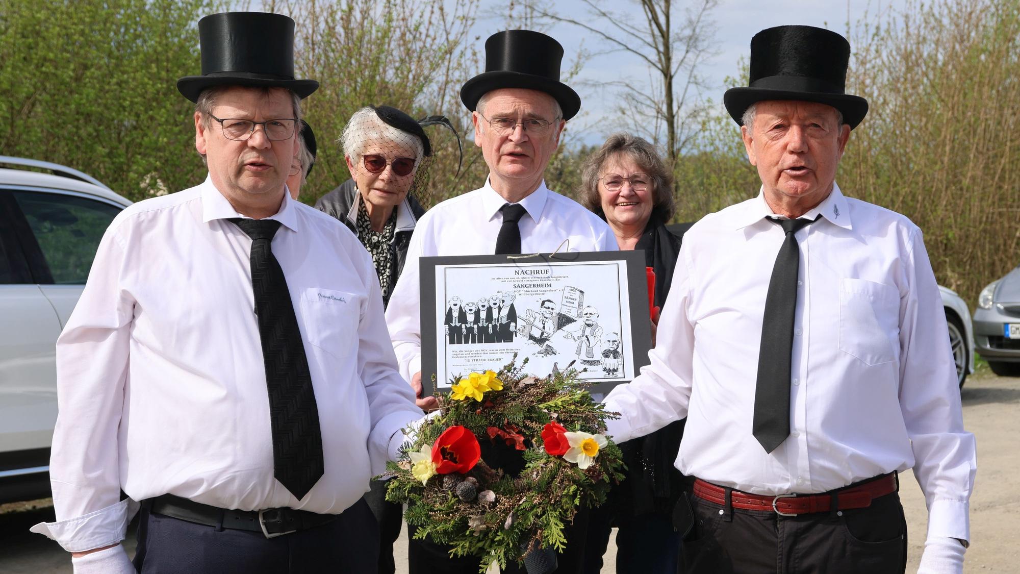 Drei Herren mit schwarzen Zylindern tragen ein Blumengesteck.