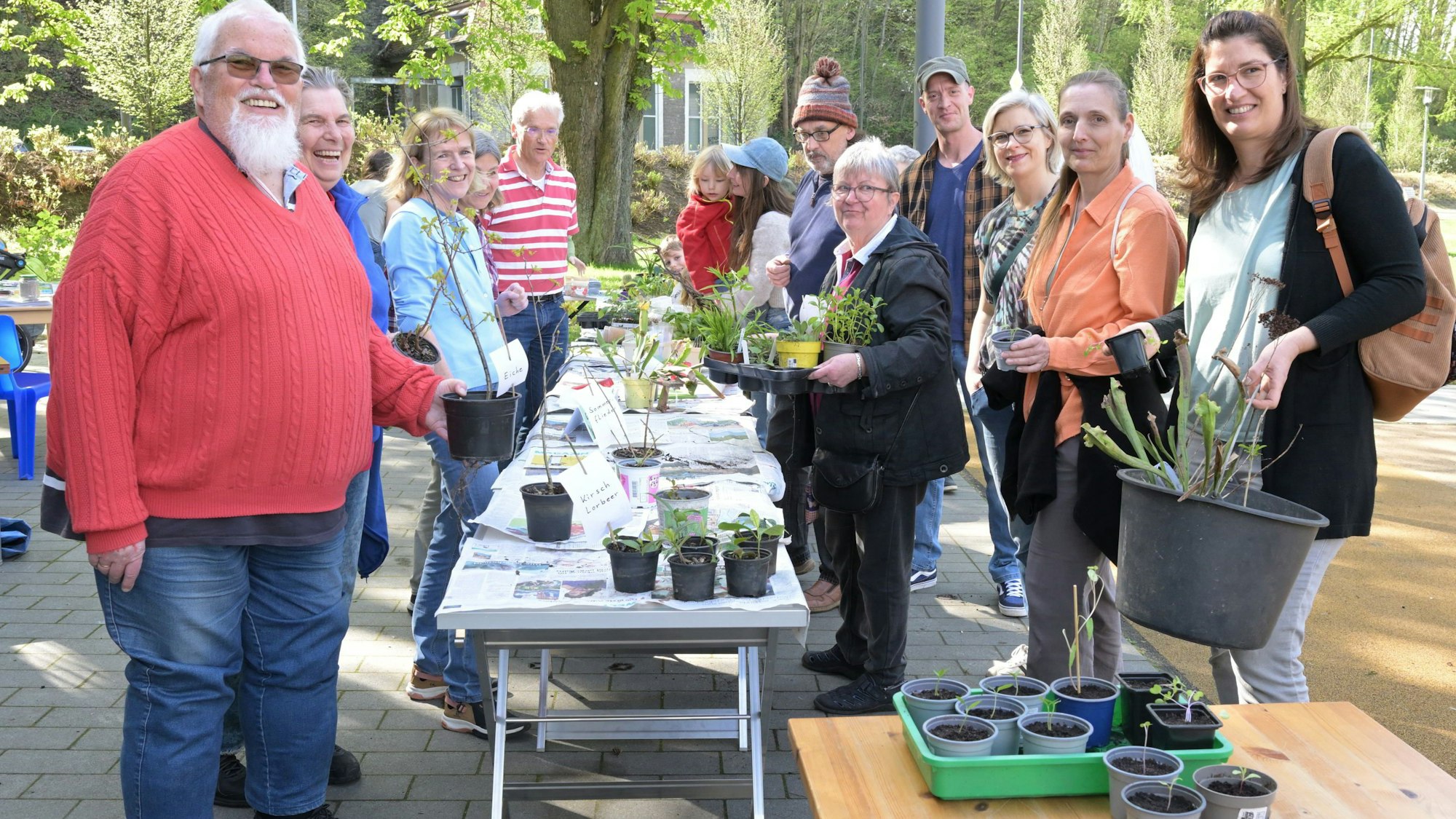 Rechts und links von einem langen Tisch stehen Männer und Frauen mit ihren kleinen und großen Pflanzen.