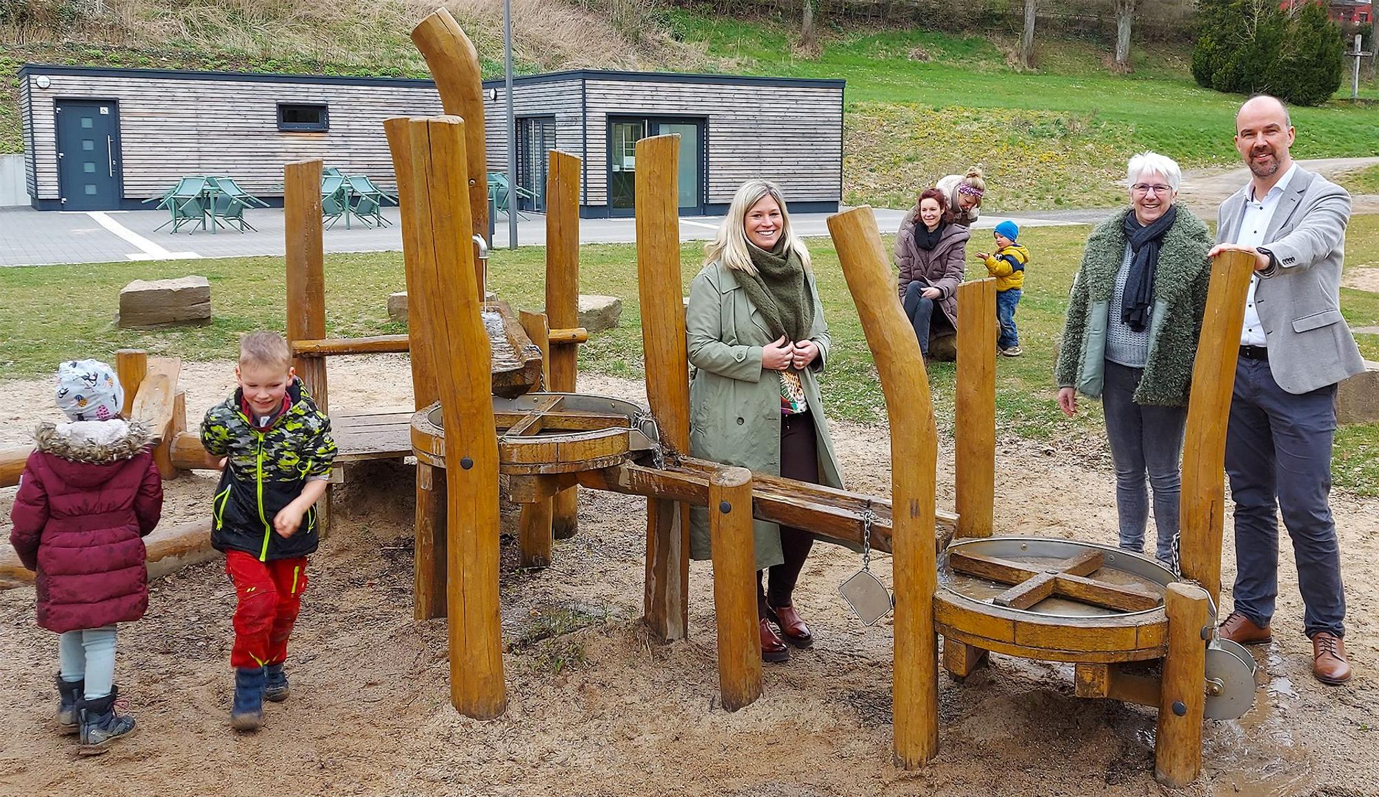 Bürgermeisterin Jennifer Meuren, Annegret Dreimüller und Guido Waters stehen am Kleinkinderspielplatz. Einige Kinder spielen.