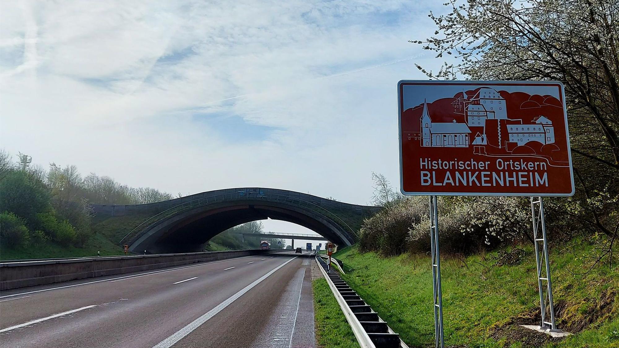 Am rechten Fahrbahnrand steht das neue Schild, auf dem zu lesen ist: „Historischer Ortskern Blankenheim“. Im Hintergrund ist die Wildbrücke zu sehen.