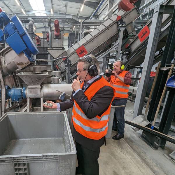 Zwei Männer in orangefarbenen Warnwesten stehen vor großen Maschinen in einer Fabrikhalle.