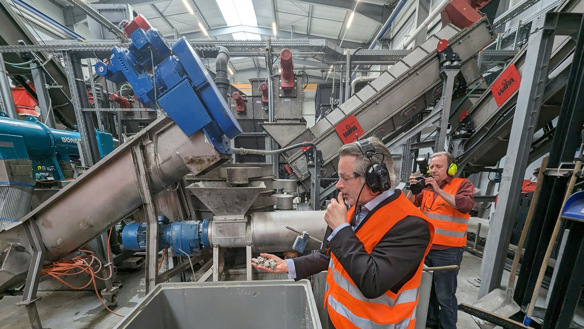 Zwei Männer in orangefarbenen Warnwesten stehen vor großen Maschinen in einer Fabrikhalle.