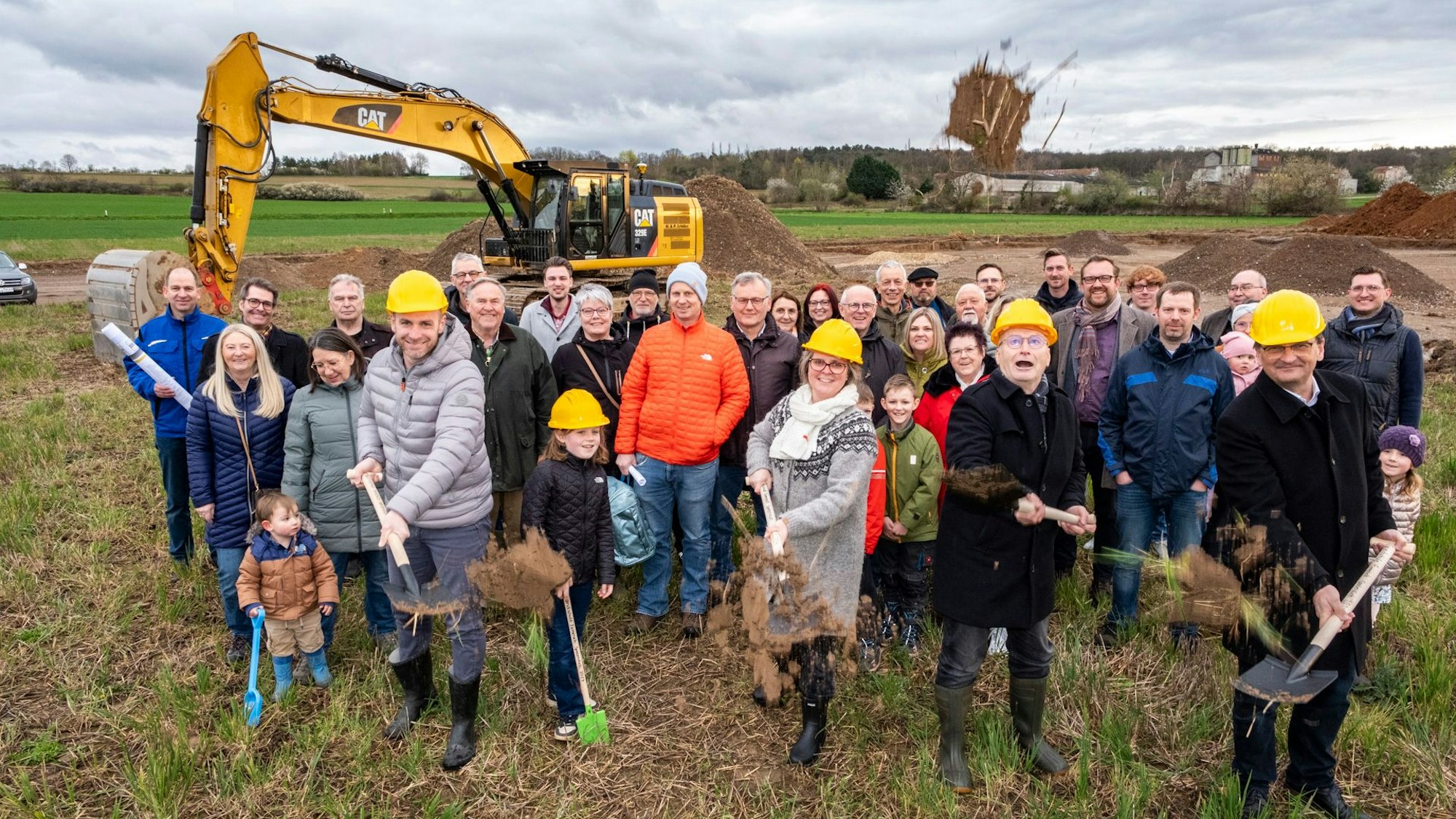 Ehrengäste des Baubeginns werfen beim ersten Spatenstich Erde in die Luft, dahinter weitere Vertreter von Politik, Verwaltung und Schulgemeinde. Auf der künftigen Baustelle steht auch schon ein Bagger.