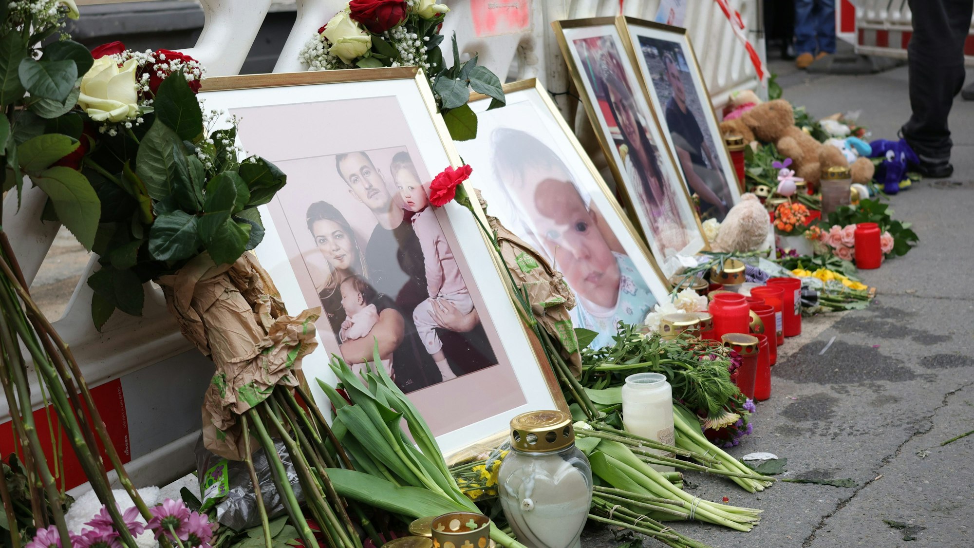 Gerahmte Fotos, Kerzen und Blumen stehen vor einer Baustellenabsperrung auf einer Straße.