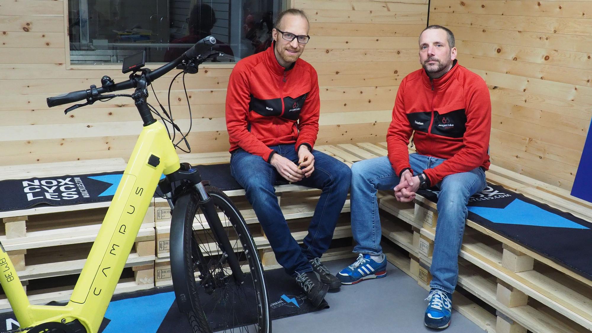 Zwei Männer sitzen auf Bänken, die aus Holz-Paletten gebaut sind. Vor ihnen steht ein gelbes Fahrrad.