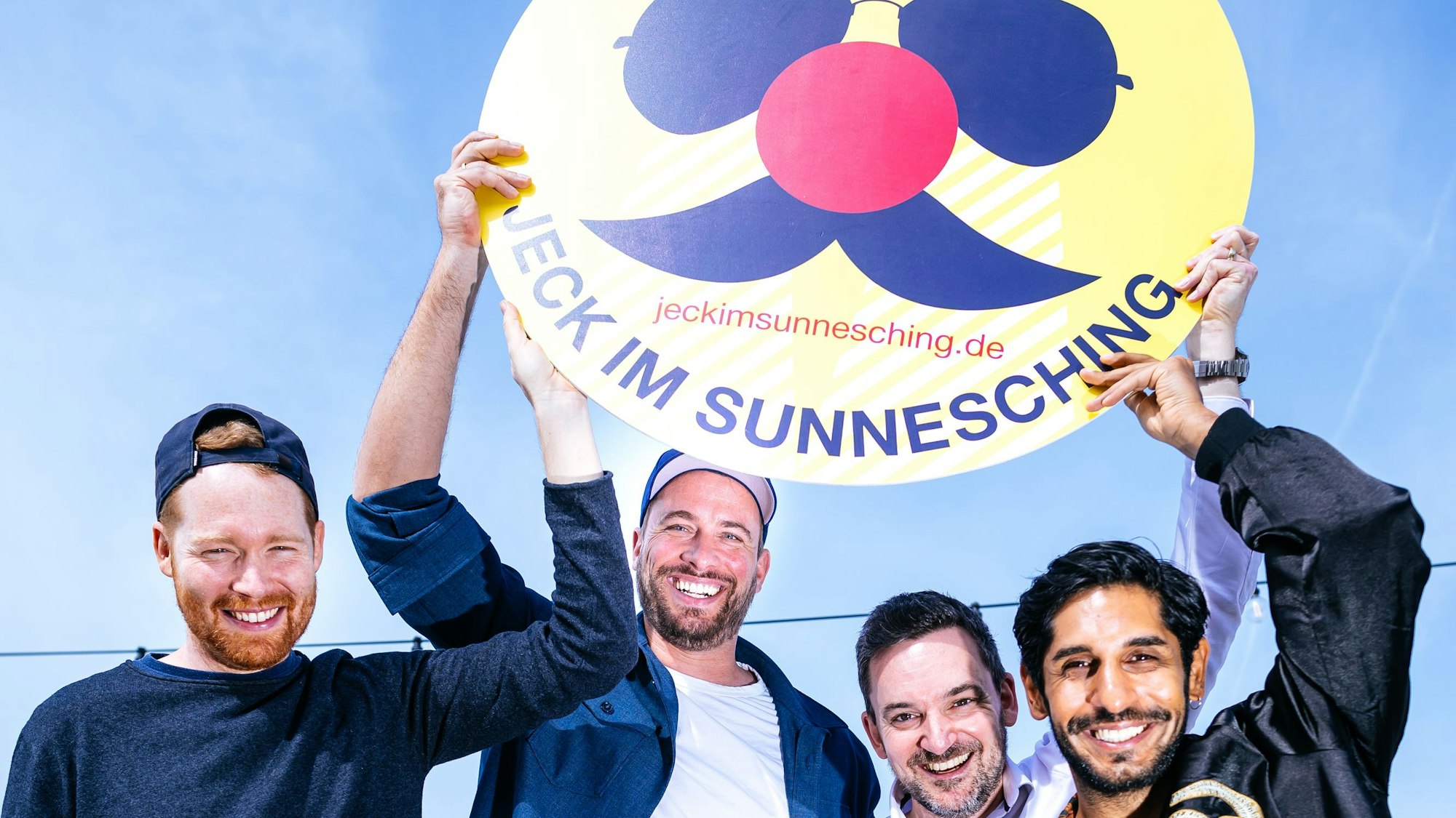 Miljö-Sänger Nils Schreiber (v.l.), Sharkys Geschäftsführer Martin Becker, Björn Lindert von x4kids und Planschemalöör-Frontmann Juri Rother halten ein Jeck im Sunnesching-Schild.