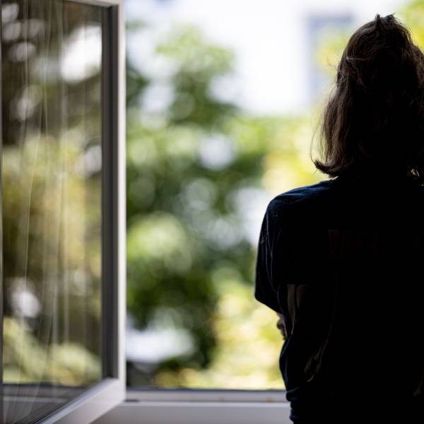 Eine Frau steht allein in ihrer Wohnung an einem Fenster und schaut ins Freie.