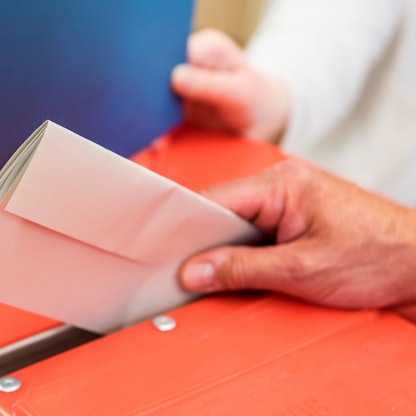 Ein Wahlhelfer steckt in einem Wahllokal den Stimmzettel eines Wählers in eine Wahlurne.