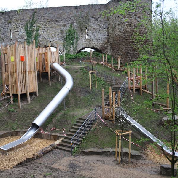 Das Bild zeigt einen Spielplatz an einem Steilhang mit zwei Rutschbahnen, einem Holzturm, einer Treppe und Sitzsteinen.